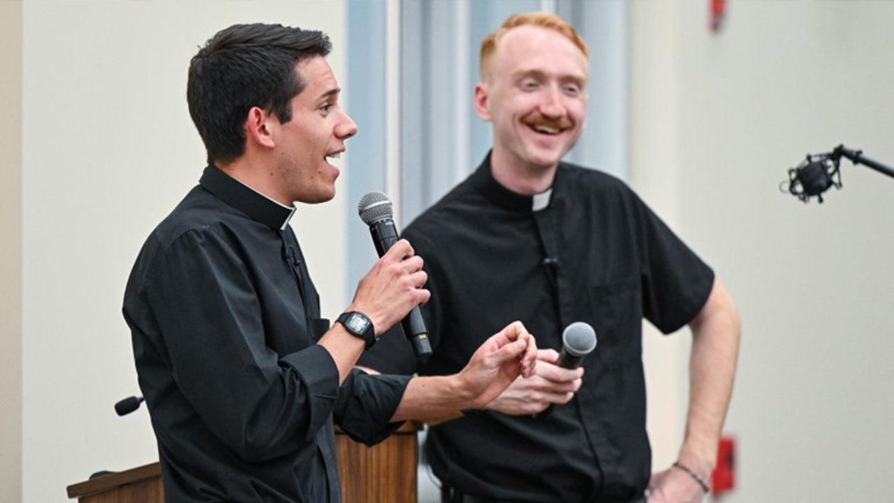 Image of Father Jeremy Leganski talking in a microphone standing next to Father Alex Lorang