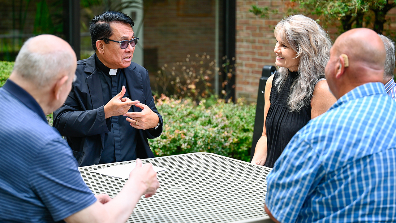 Five people looking at a priest all sitting at a table outside